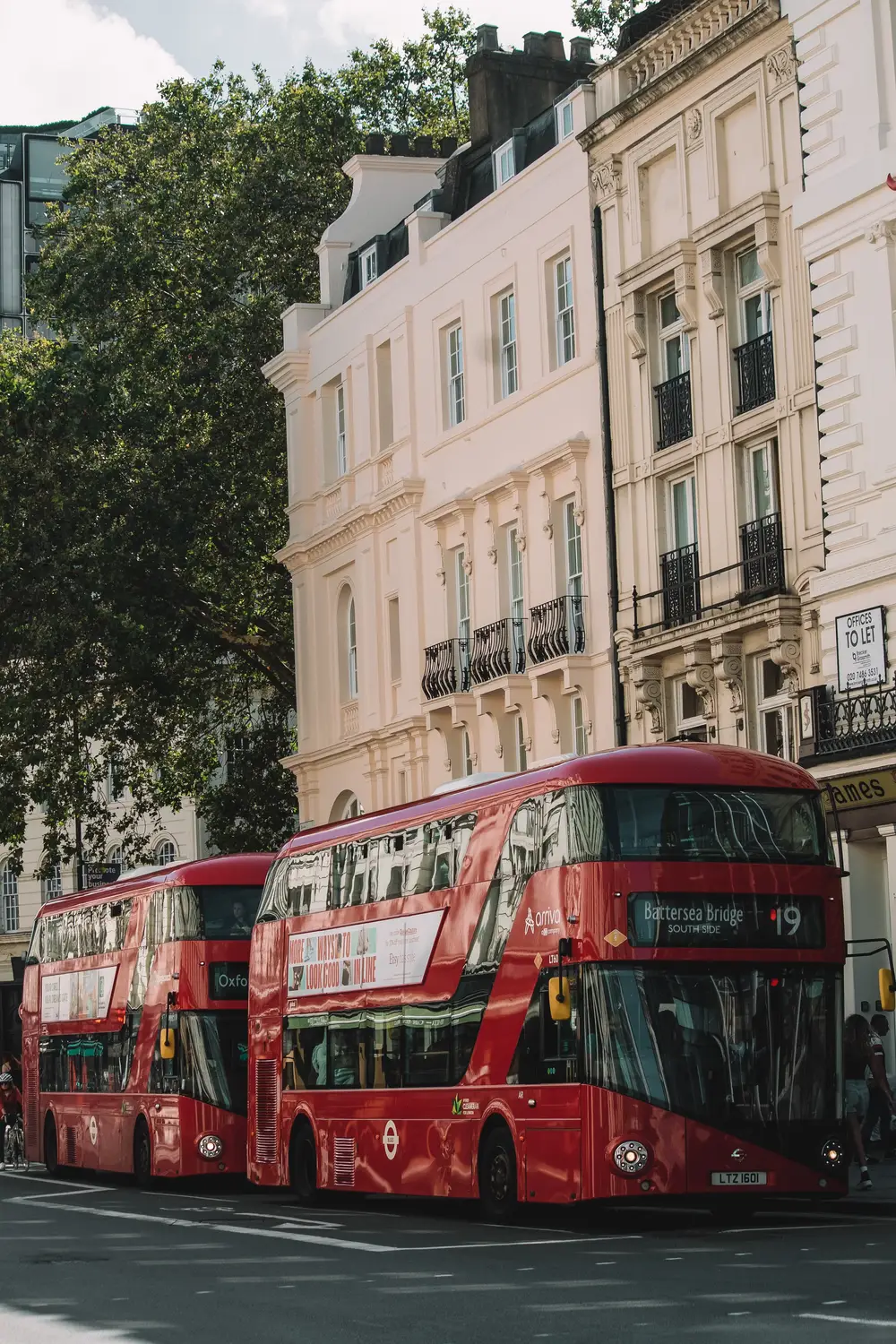 Red double-decker buses driving past elegant white period buildings on a tree-lined London street.