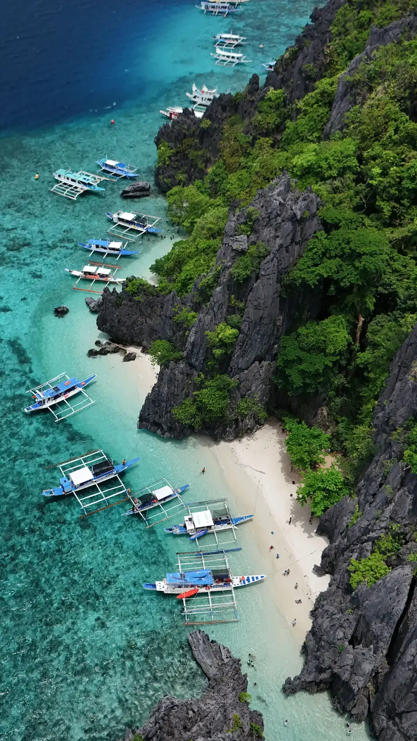 Aerial view of traditional outrigger boats anchored along a white sand beach with turquoise water and limestone cliffs in El Nido, Palawan, Philippines.