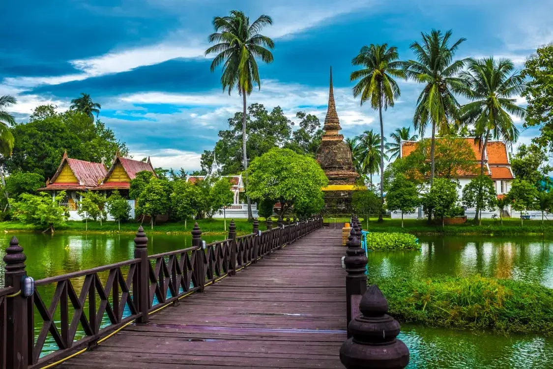  Wooden bridge over a pond leading to an ancient brick stupa amid palm trees and Thai temple buildings.