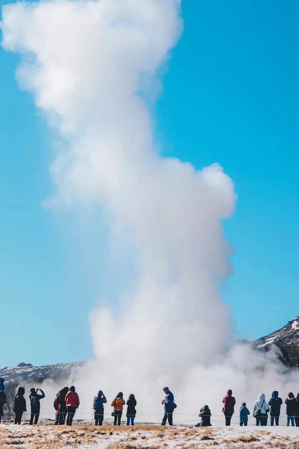 Crowd watching a geyser erupt into the sky under clear blue weather, Iceland.