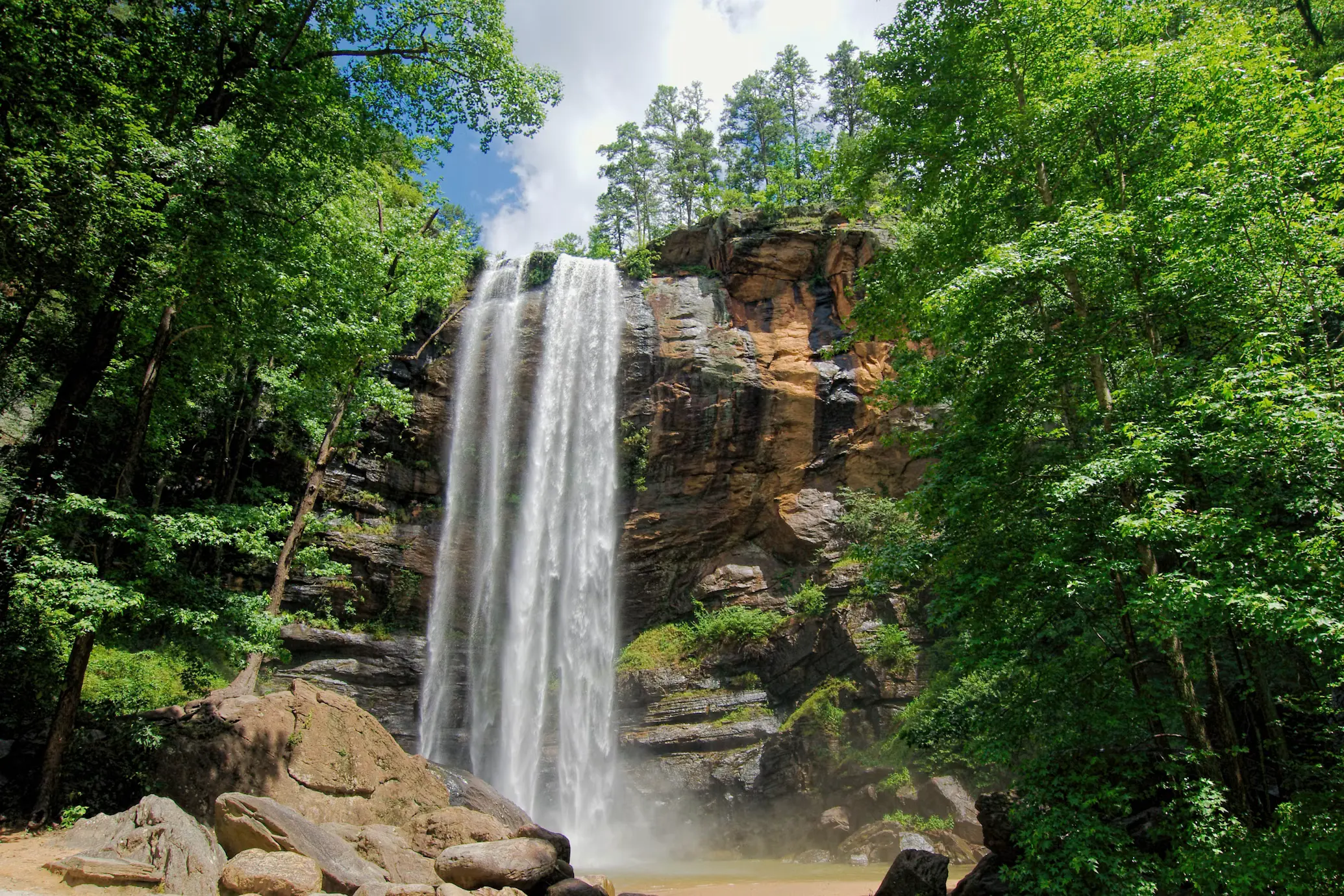 Tall waterfall cascading over a rocky cliff into a pool, surrounded by lush green forest.