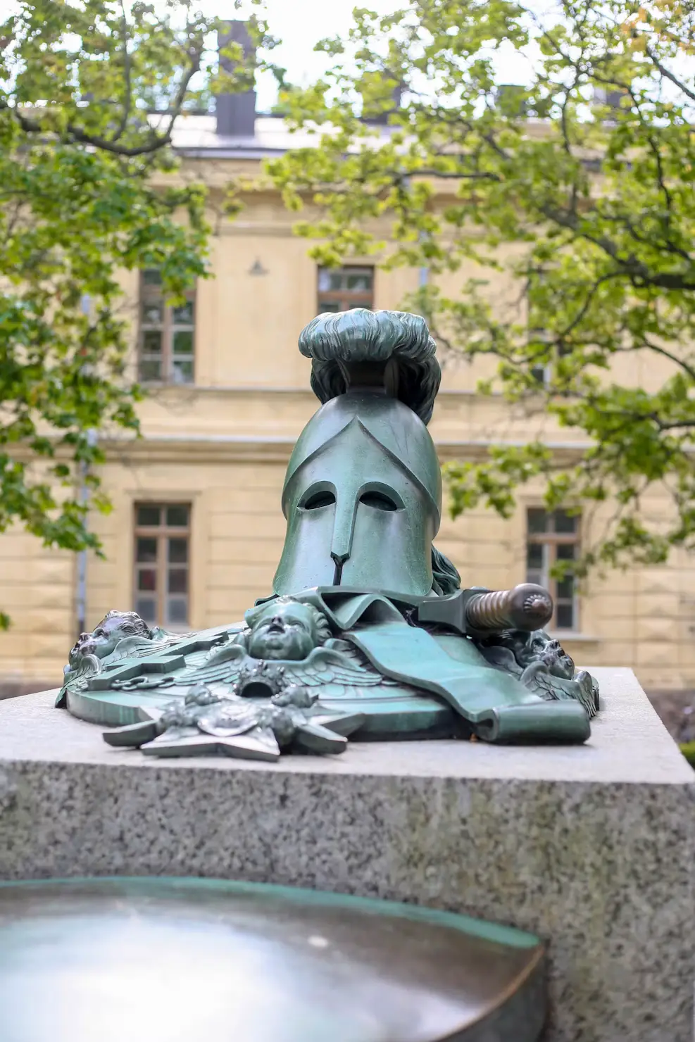 Bronze sculpture of a classical helmet, sword, and decorative figures on a stone pedestal in front of a historic building and leafy trees.