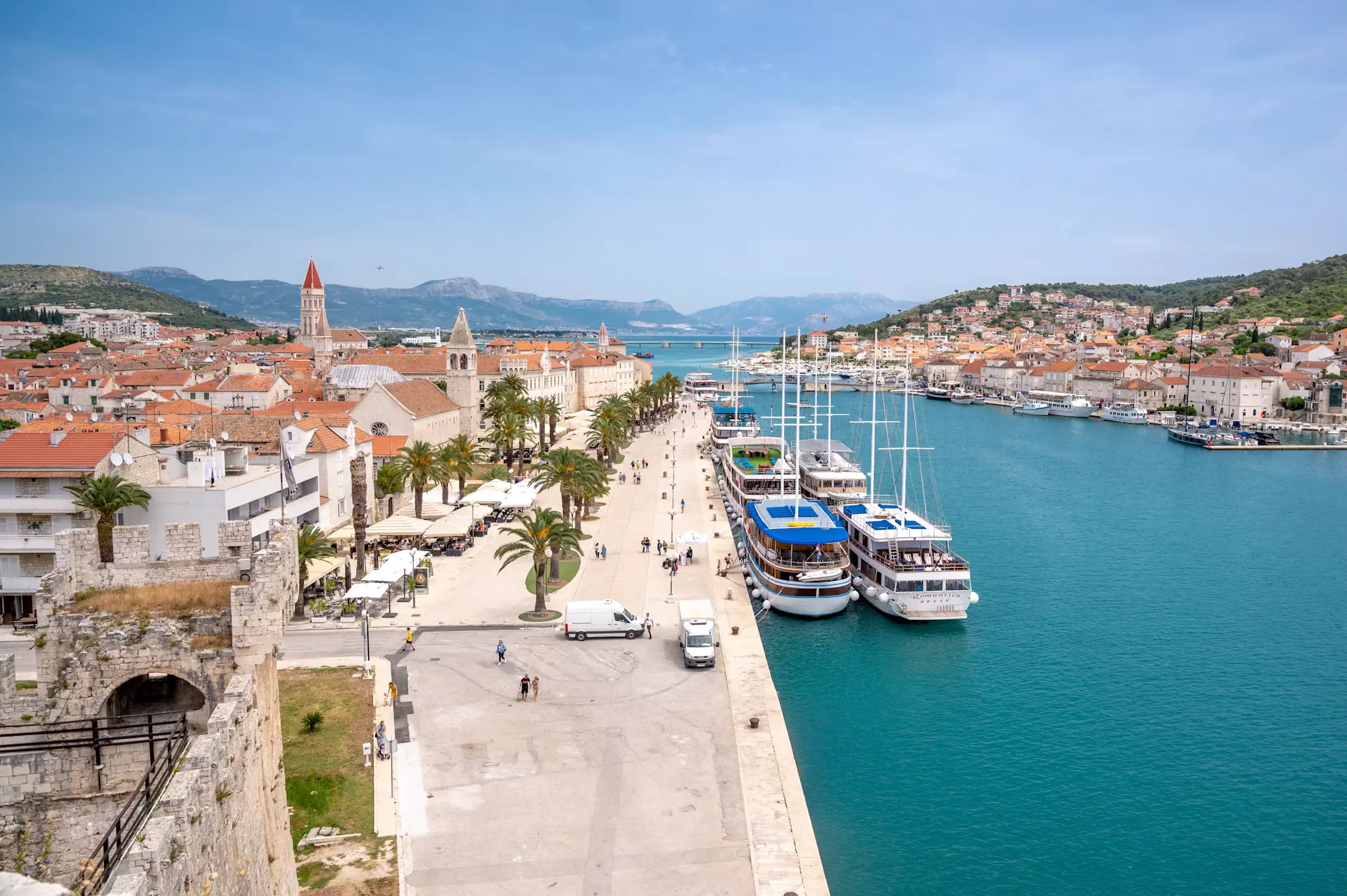 Aerial view of Trogir's stone waterfront promenade lined with palm trees and yachts, historic town buildings and green hills in the background, Croatia.