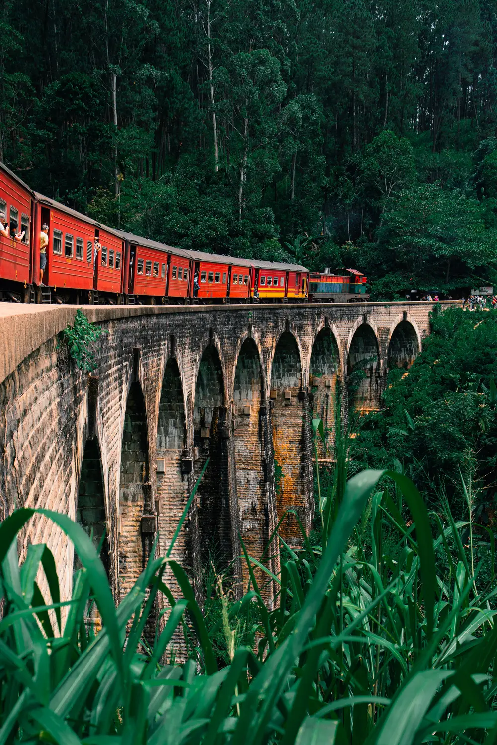 Red train crossing the Nine Arch Bridge surrounded by lush green jungle in Ella, Sri Lanka.