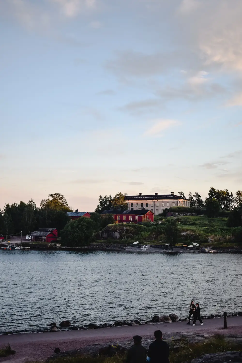 Waterfront at dusk with red houses on a green hillside, calm water, and people walking along a rocky shoreline path.