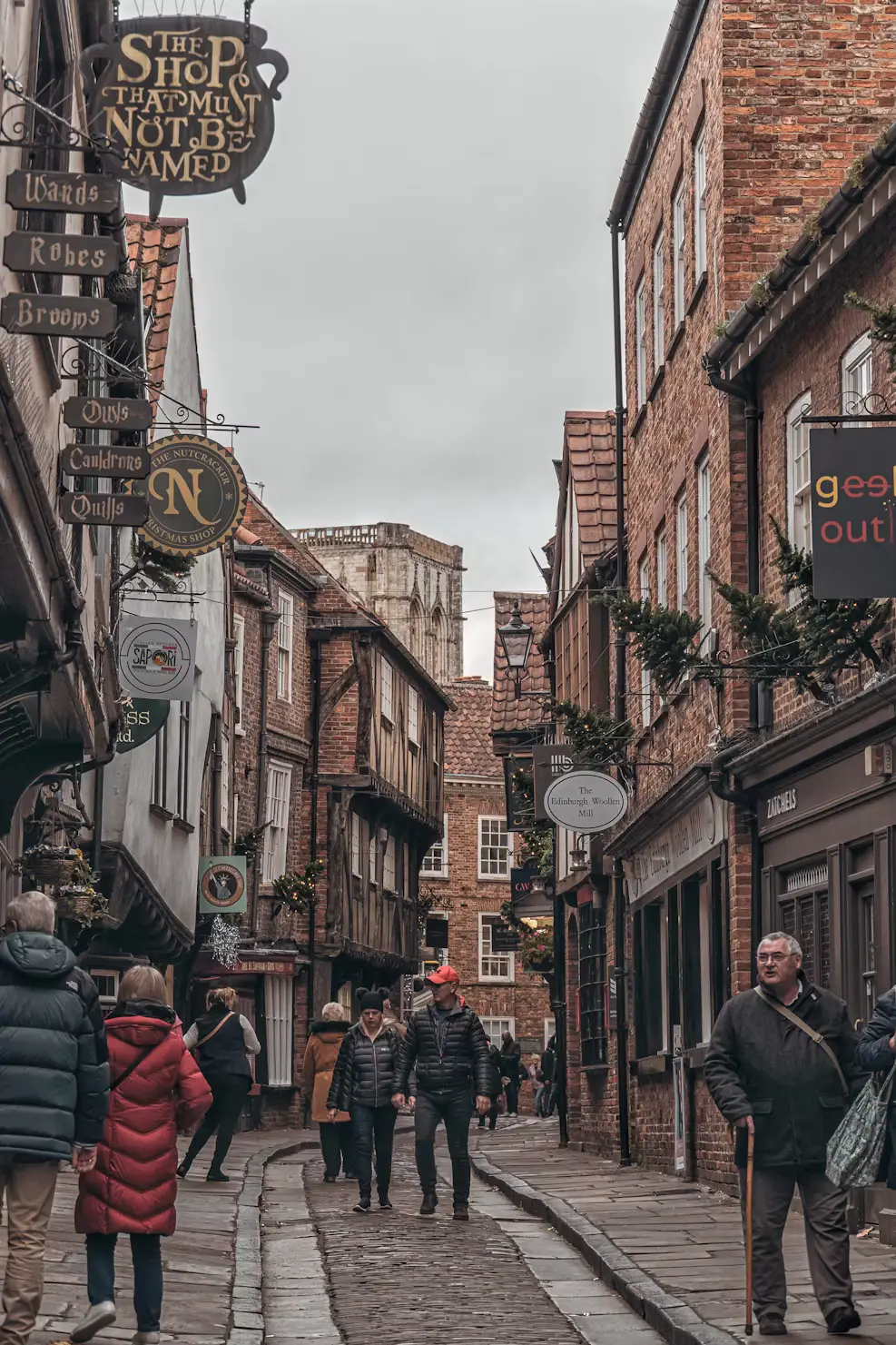 Pedestrians strolling down the Shambles in York, a narrow cobbled lane lined with crooked medieval buildings and hanging shop signs.