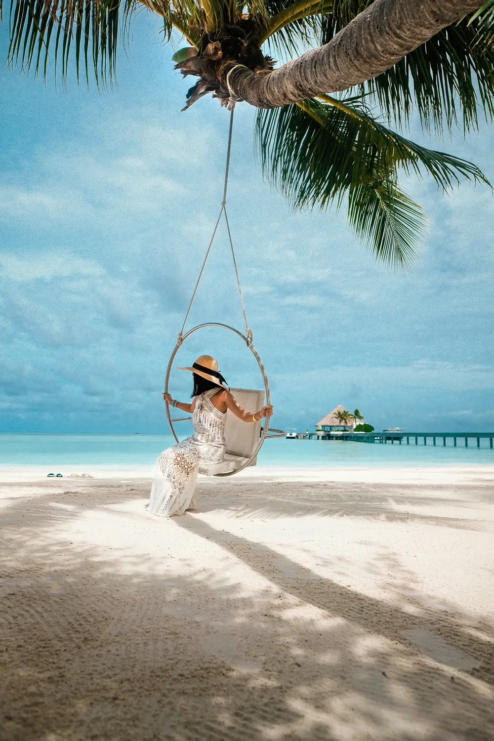 Woman sitting on palm tree rope swing on white sand beach with overwater bungalow jetty and turquoise lagoon in Maldives