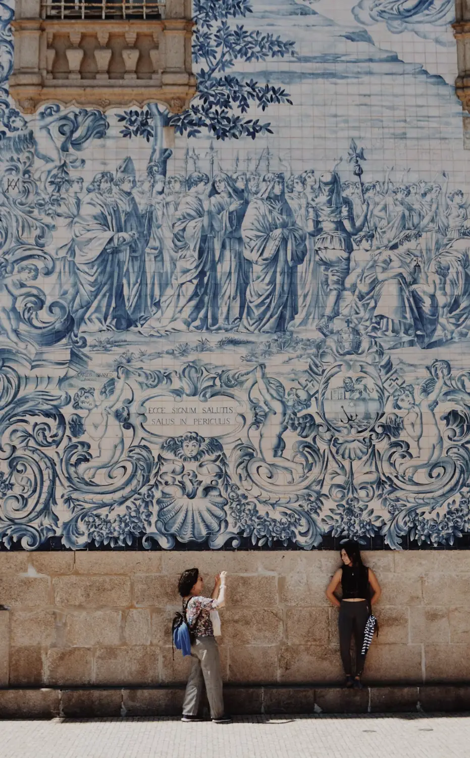 Two people stand beneath a large blue-and-white azulejo mural depicting a historical procession on a stone building wall.