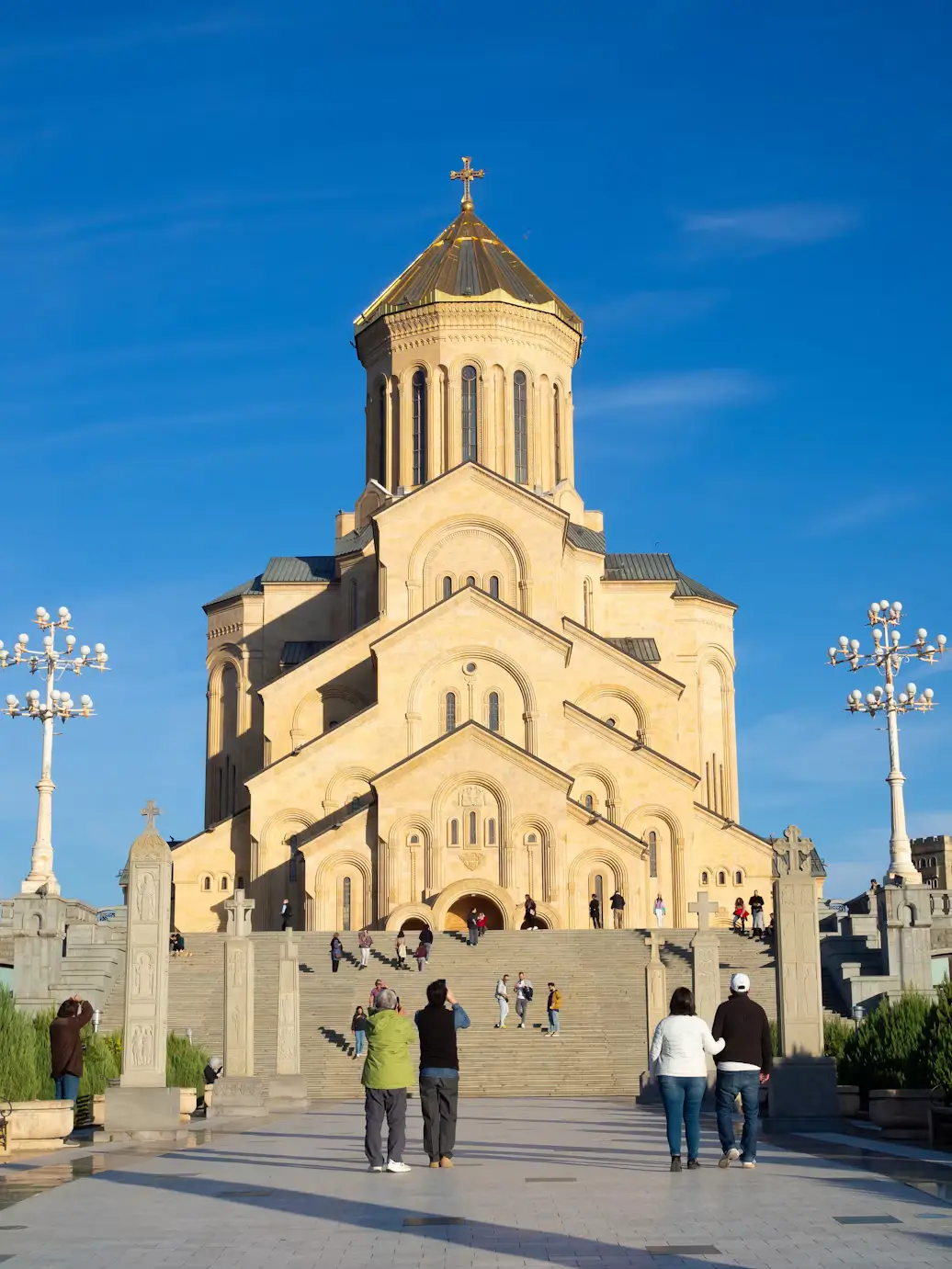 A large ornate cathedral with a golden dome and cross, viewed from a broad plaza under a bright blue sky.