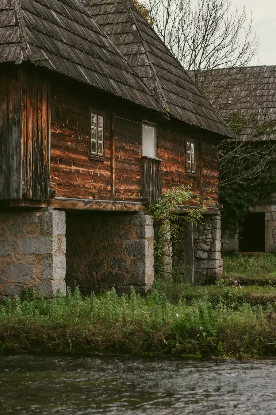 Old wooden watermill house on stilts beside a stream, surrounded by greenery.