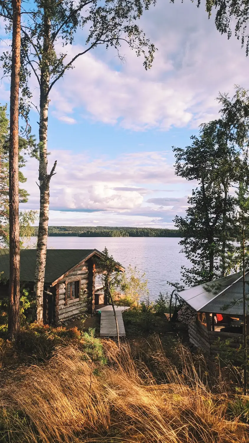 Lakeside log cabin surrounded by tall trees under a partly cloudy sky.