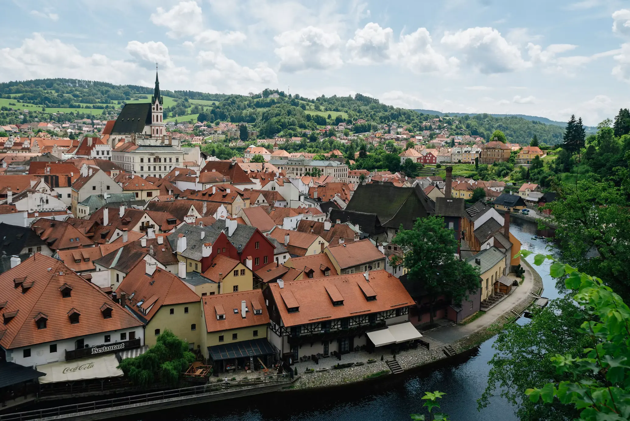 Aerial view of Český Krumlov's historic old town with red-roofed buildings along a bend in the Vltava River, surrounded by green hills