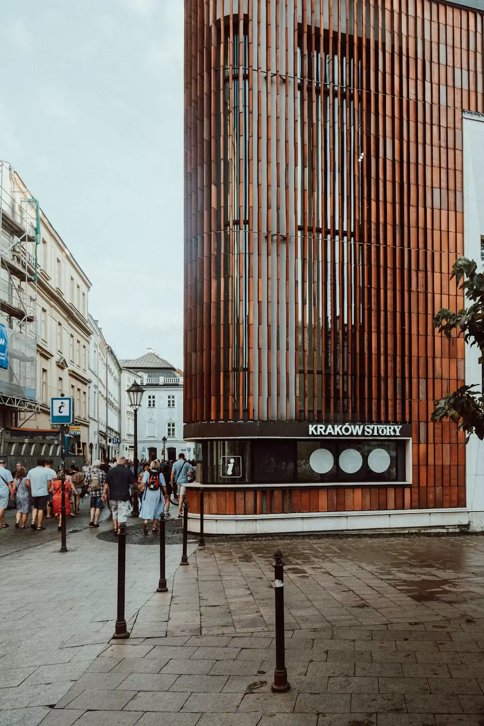 A street scene in Kraków, Poland, featuring a modern terracotta-clad building, historic architecture, pedestrians, and a “KRAKÓW STORY” information kiosk.