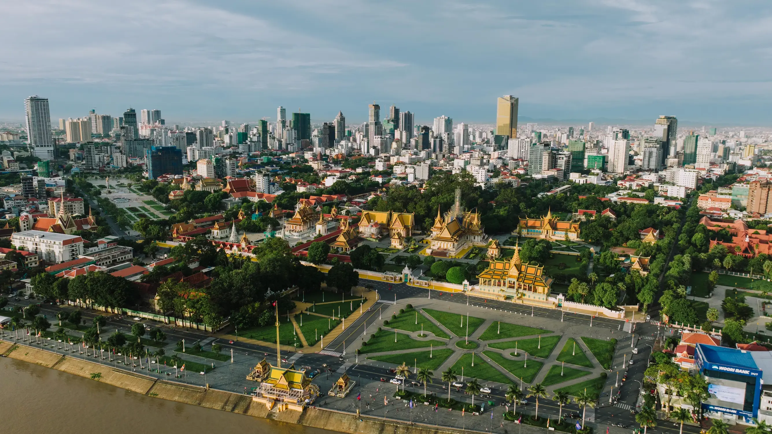 Aerial drone view of Royal Palace complex with golden temples and green gardens along Tonle Sap riverfront and Phnom Penh city skyline, Cambodia