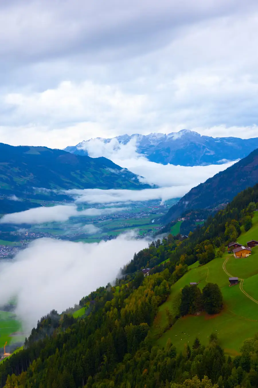 A misty valley filled with low clouds beneath forested mountains, with green pastures and scattered farmhouses on the hillside below.
