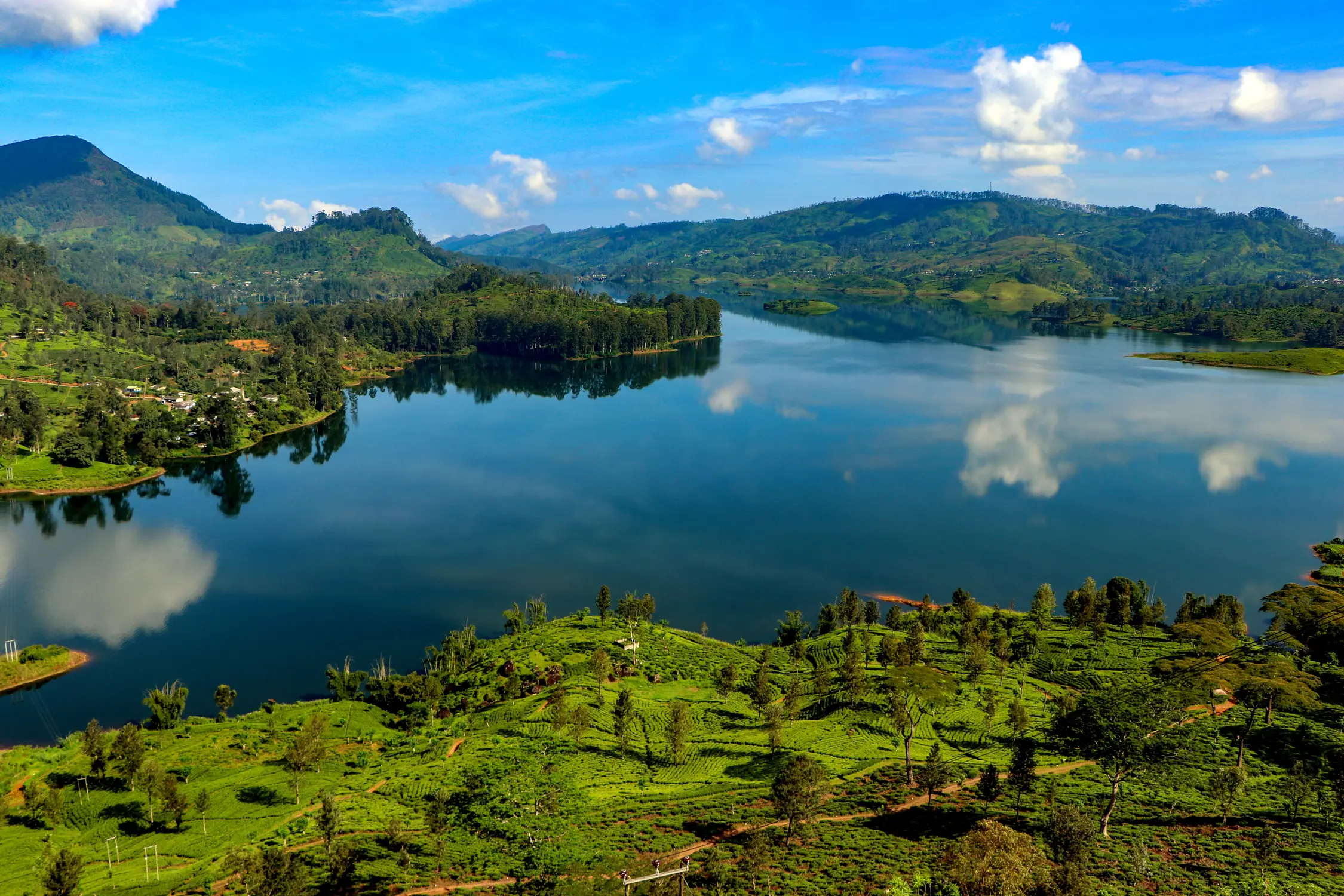 Calm lake reflecting blue sky and clouds, surrounded by lush green hills and islands in Sri Lanka.