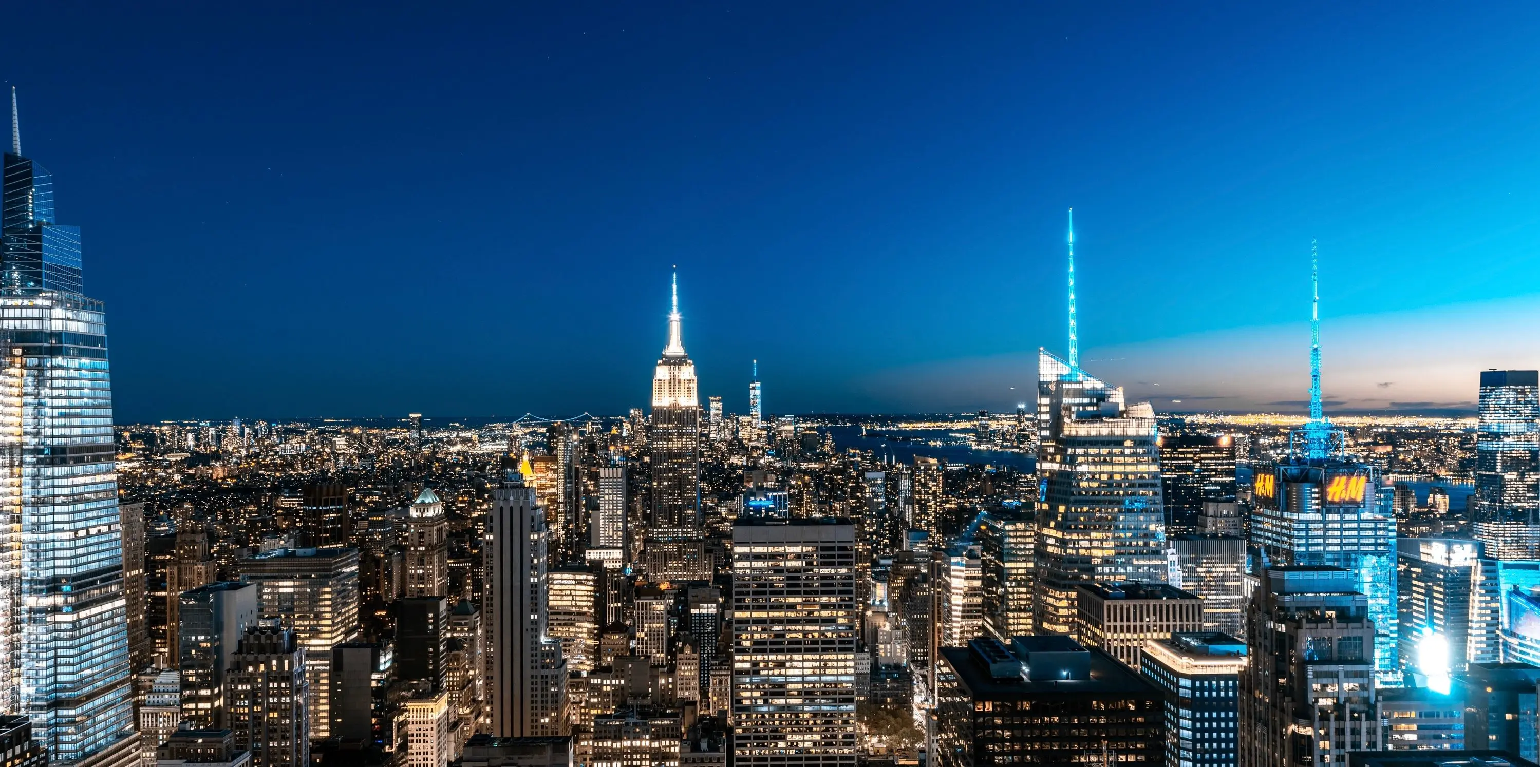 Panoramic view of the Manhattan skyline at dusk, with the Empire State Building and illuminated skyscraper spires glowing against a deep blue sky.