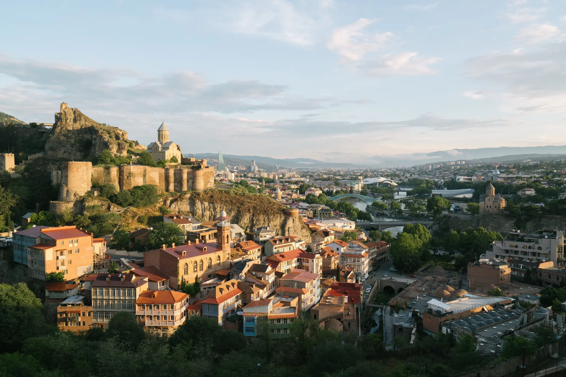 A panoramic view of Tbilisi, Georgia, showing the historic Narikala Fortress and church on a rocky hillside, red-roofed buildings in the foreground, the modern city and river bridges beyond, and rolling hills under a partly cloudy sky.