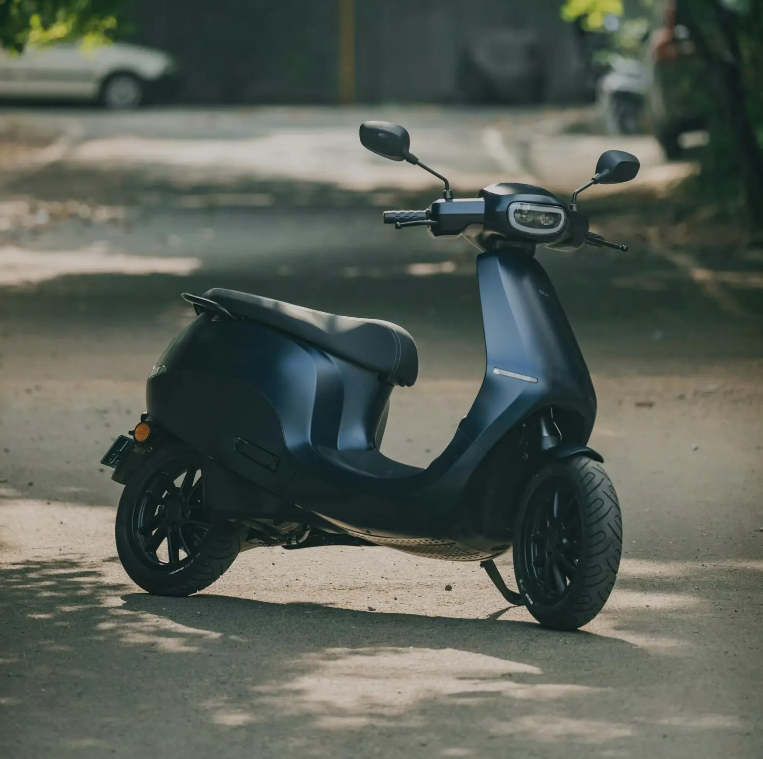 A matte black electric scooter parked on a quiet tree-lined street, with dappled sunlight filtering through green leaves onto the pavement