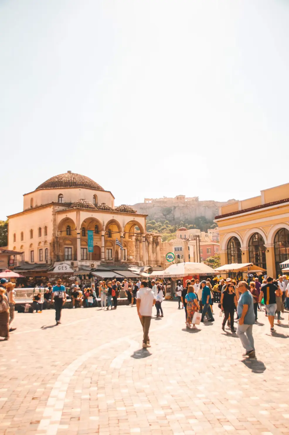 A bustling square in Athens with a historic mosque building, market stalls, and pedestrians, with the Acropolis visible in the background.