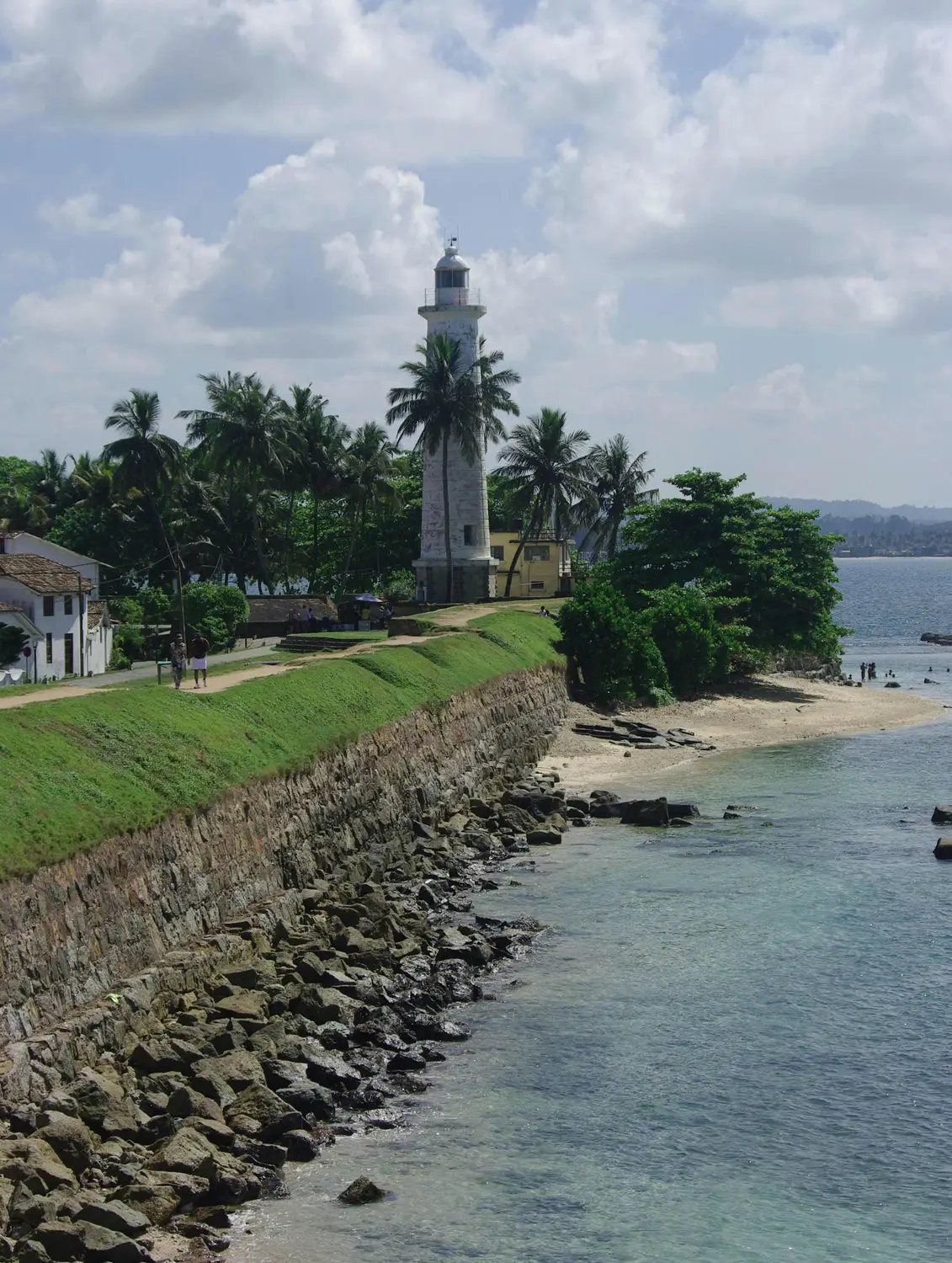 White Galle Lighthouse surrounded by palm trees along the historic fort ramparts and rocky coastline in Galle, Sri Lanka.