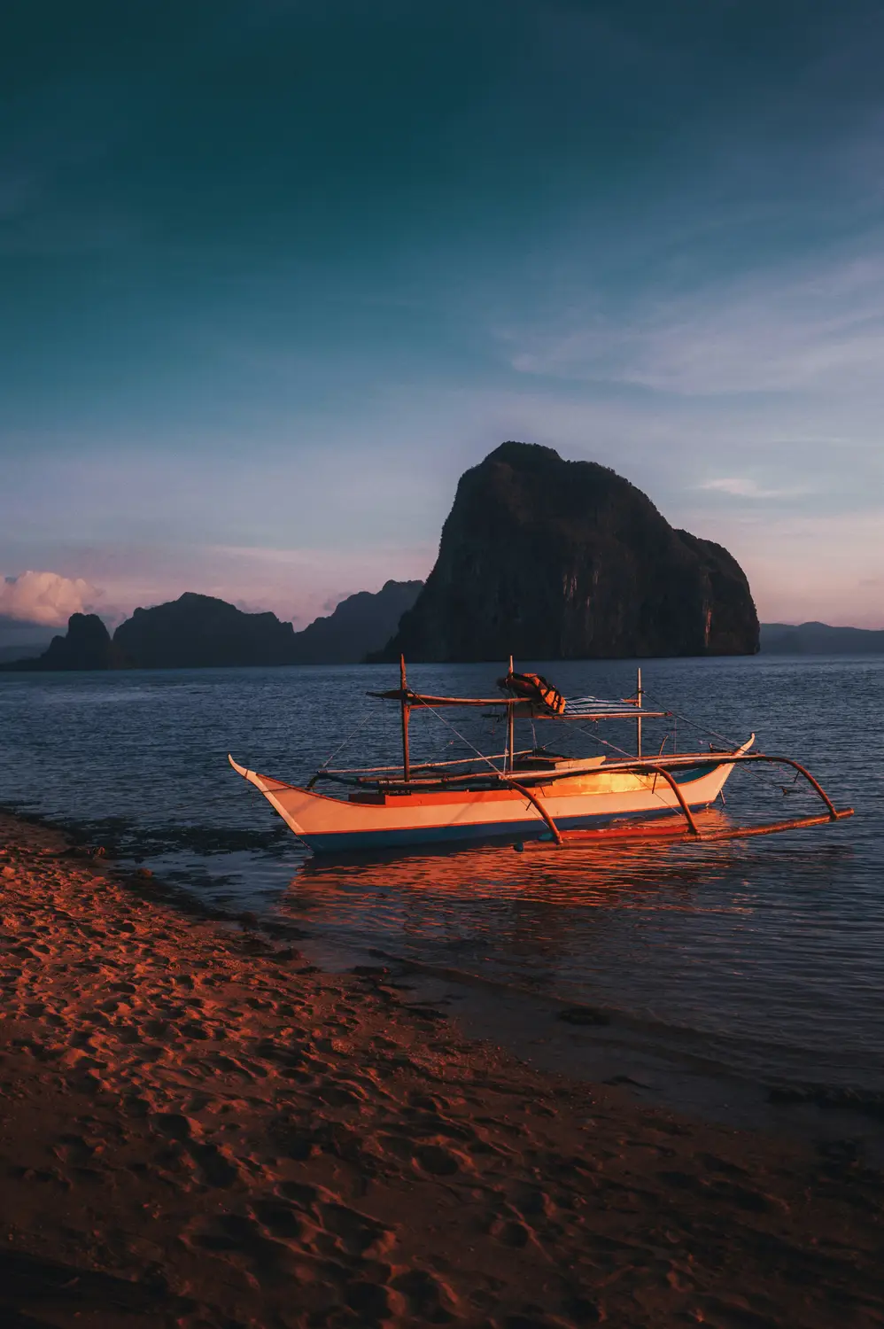 Traditional orange outrigger boat on a beach at dusk with karst islands in El Nido, Palawan, Philippines.