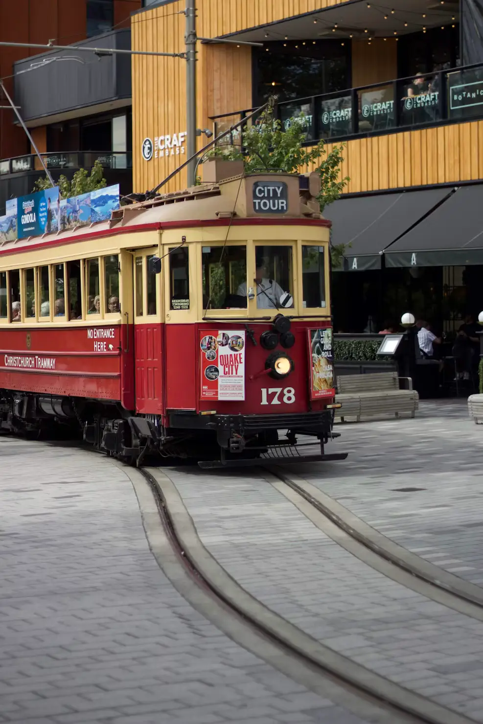 Vintage red Christchurch tram traveling through a modern city street with shops and outdoor seating.