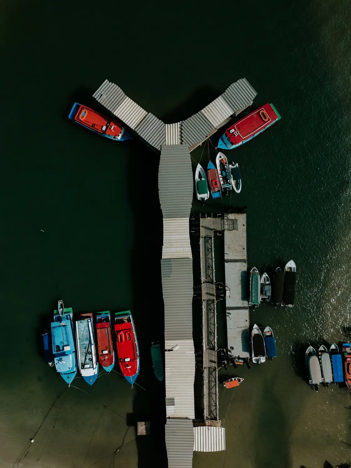 Aerial drone view of colourful boats moored at Y-shaped wooden jetty pier on green water