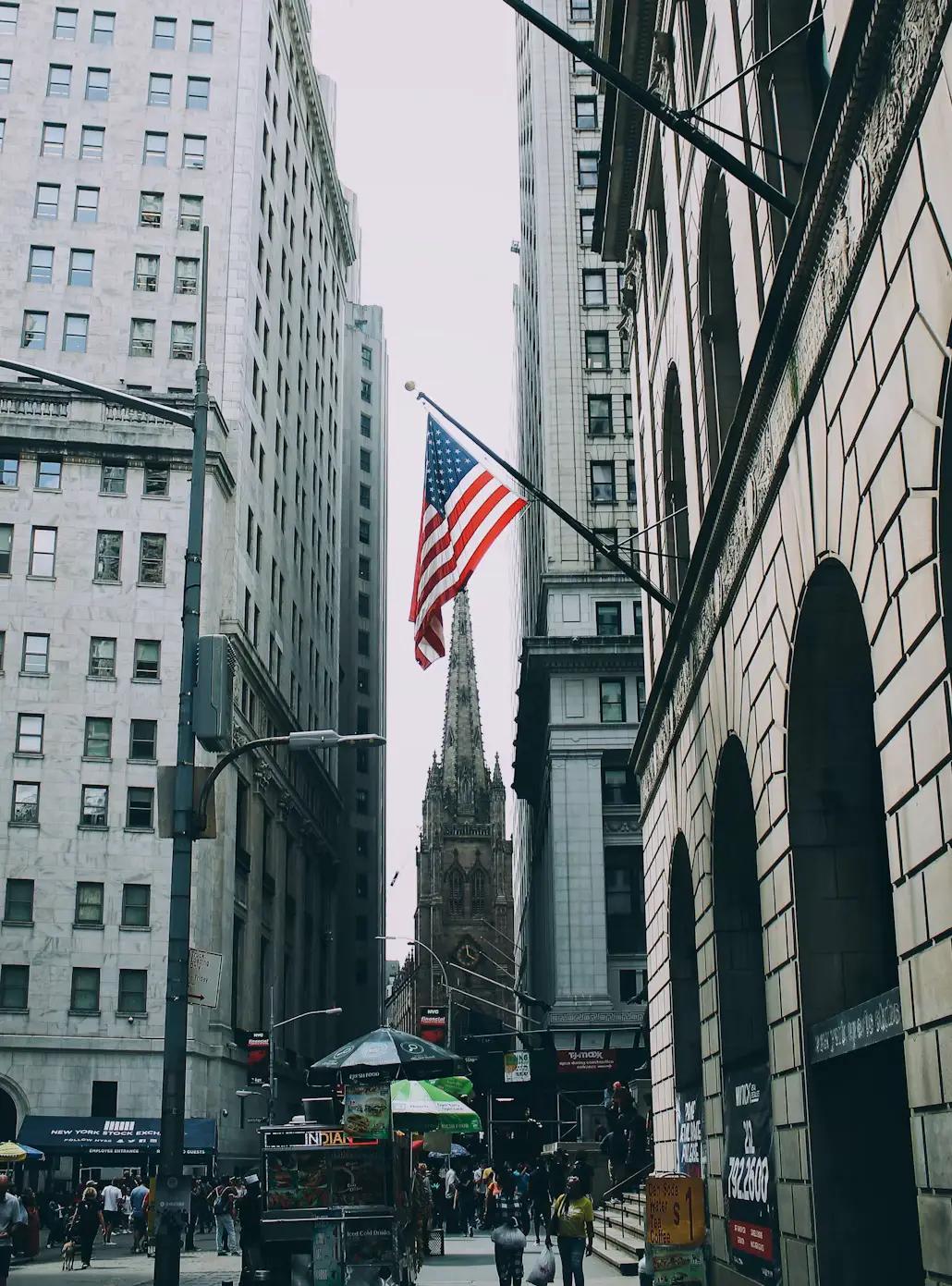 An American flag flying over a busy Lower Manhattan street, with a gothic church spire framed between tall stone buildings.