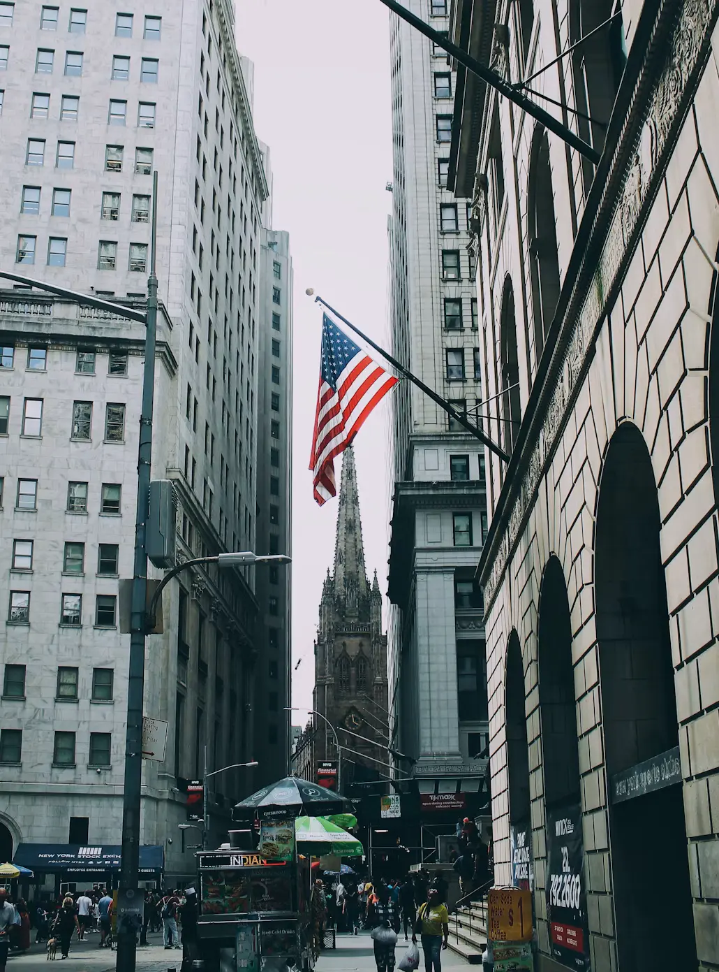 An American flag flying over a busy Lower Manhattan street, with a gothic church spire framed between tall stone buildings.