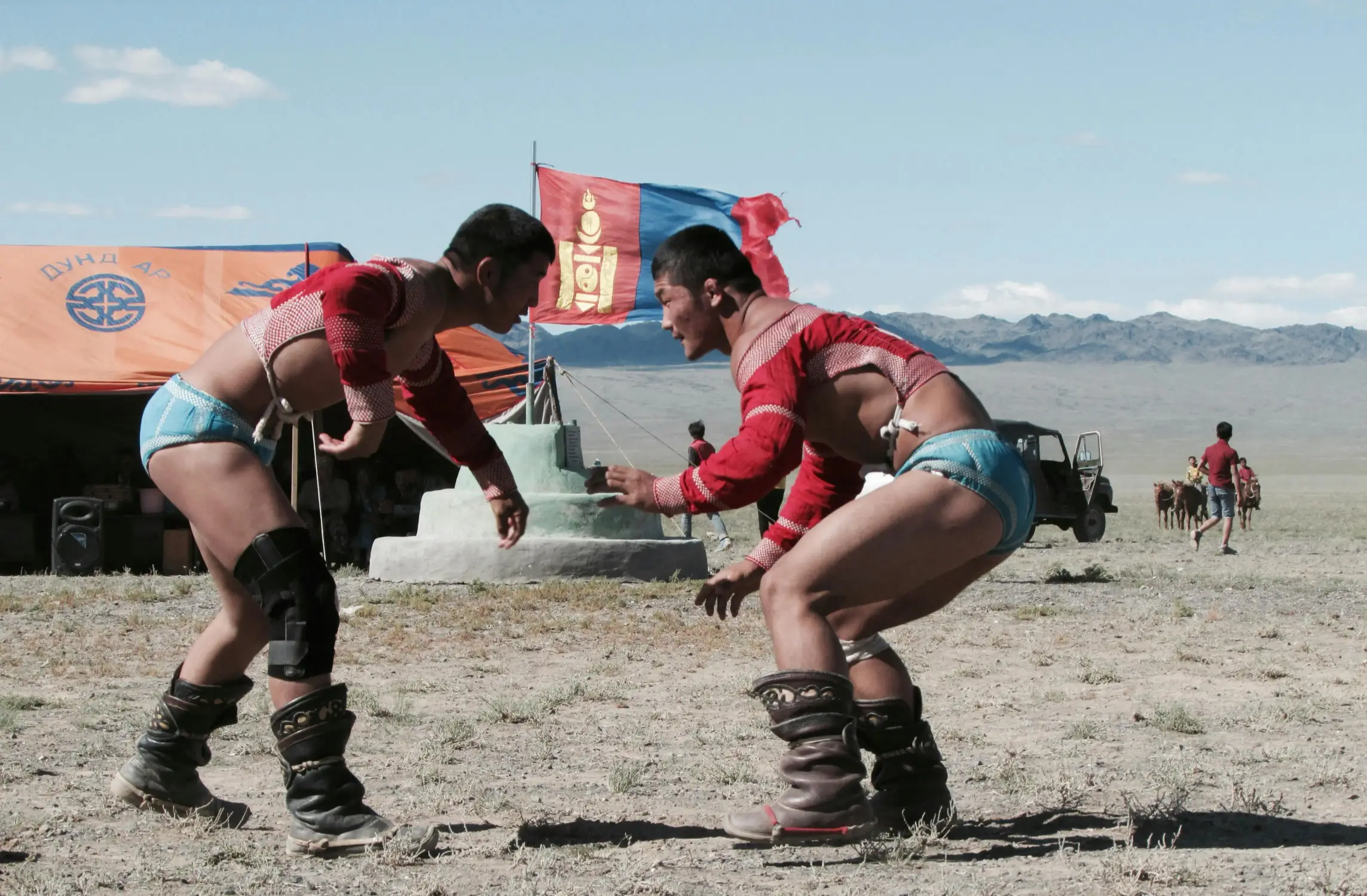 Two Mongolian wrestlers in traditional costume competing at an outdoor Naadam festival, with the Mongolian flag and vast steppe landscape in the background.