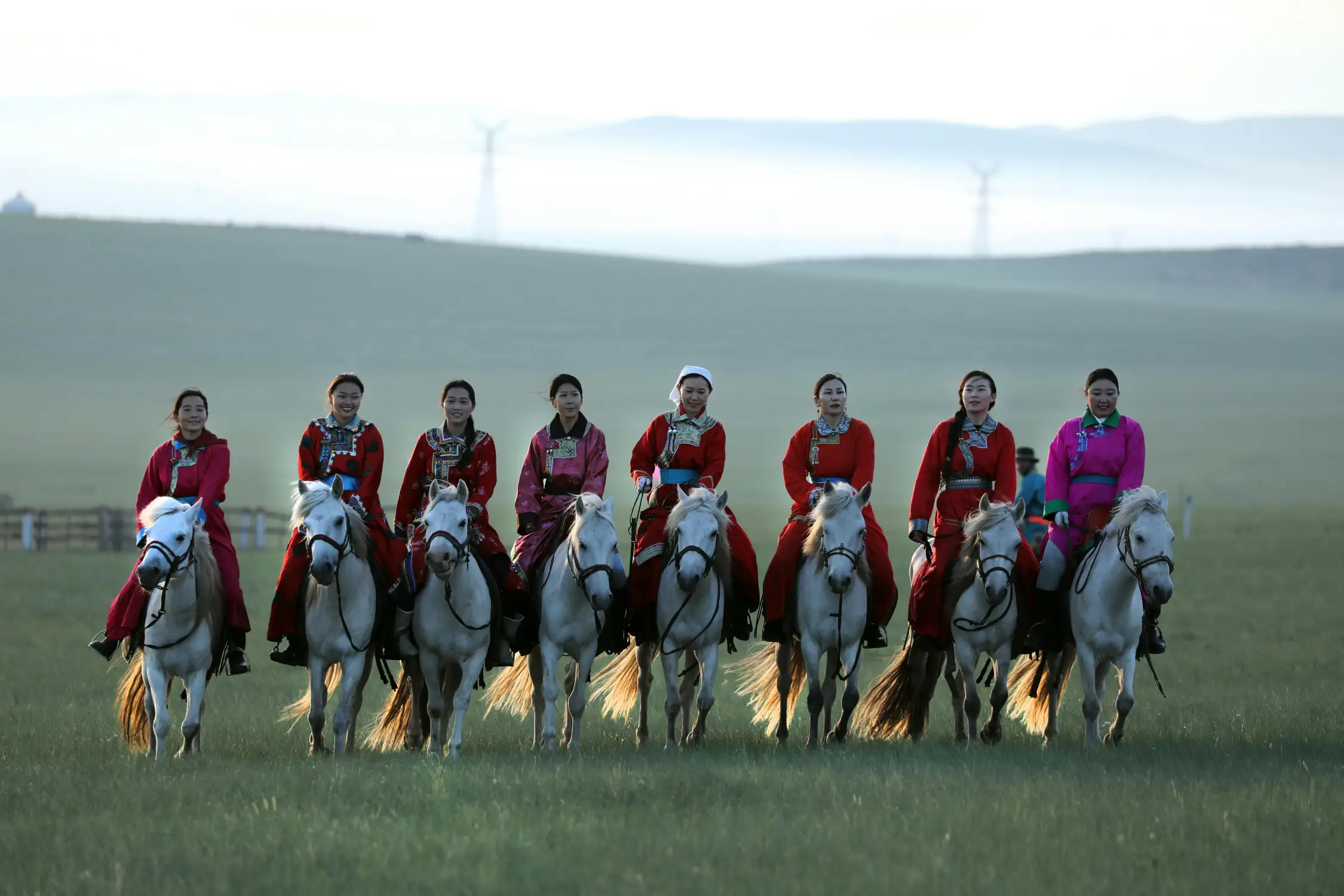 Group of Mongolian women in vibrant red traditional deel costumes riding white horses across the green steppe at Naadam Festival, Mongolia
