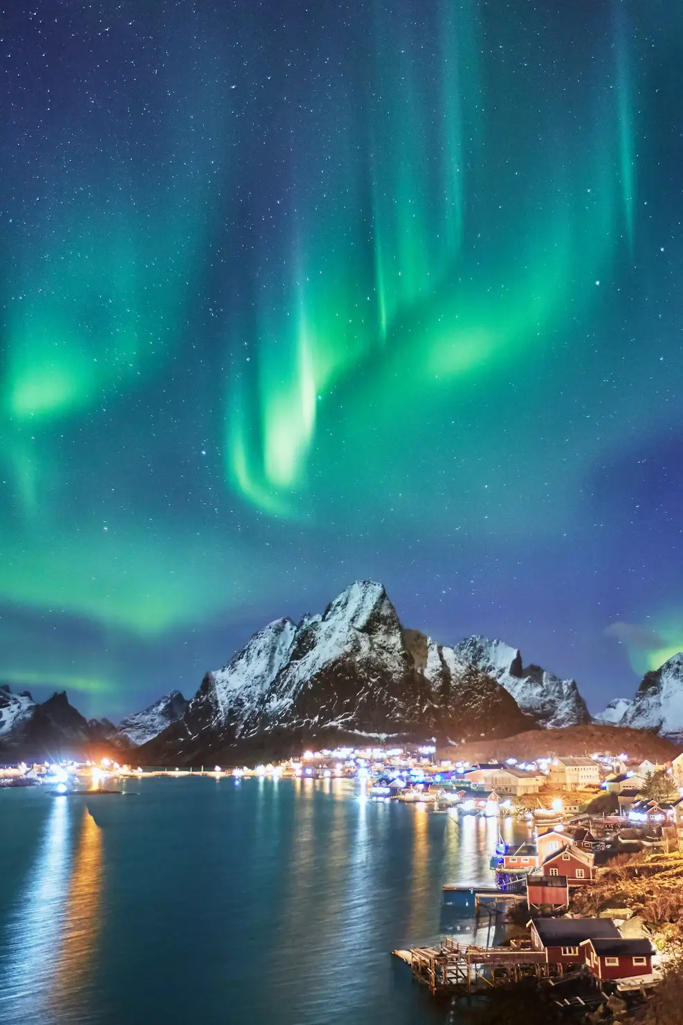 Green northern lights swirling in the night sky above a snow-capped mountain and the glowing lights of a coastal village.