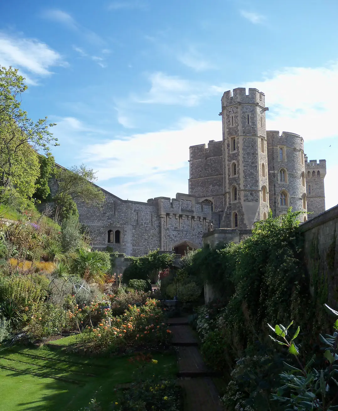 Stone towers of Windsor Castle rising above a lush terraced garden filled with shrubs and flowers under a bright blue sky.
