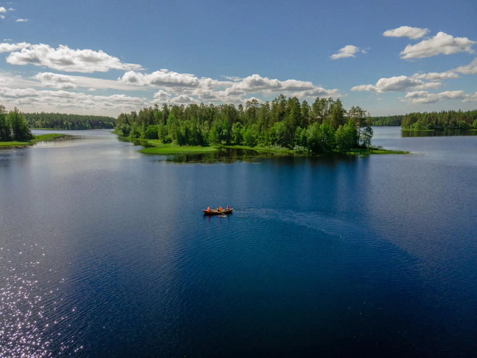 Small boat crossing a calm blue lake with a forested island under a bright, cloud-filled sky.