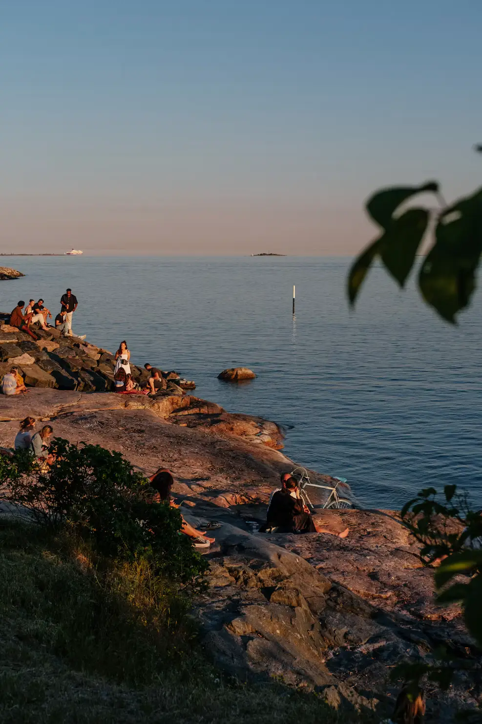 People relaxing on a rocky shoreline by calm water at sunset, with distant islands and a boat on the horizon.