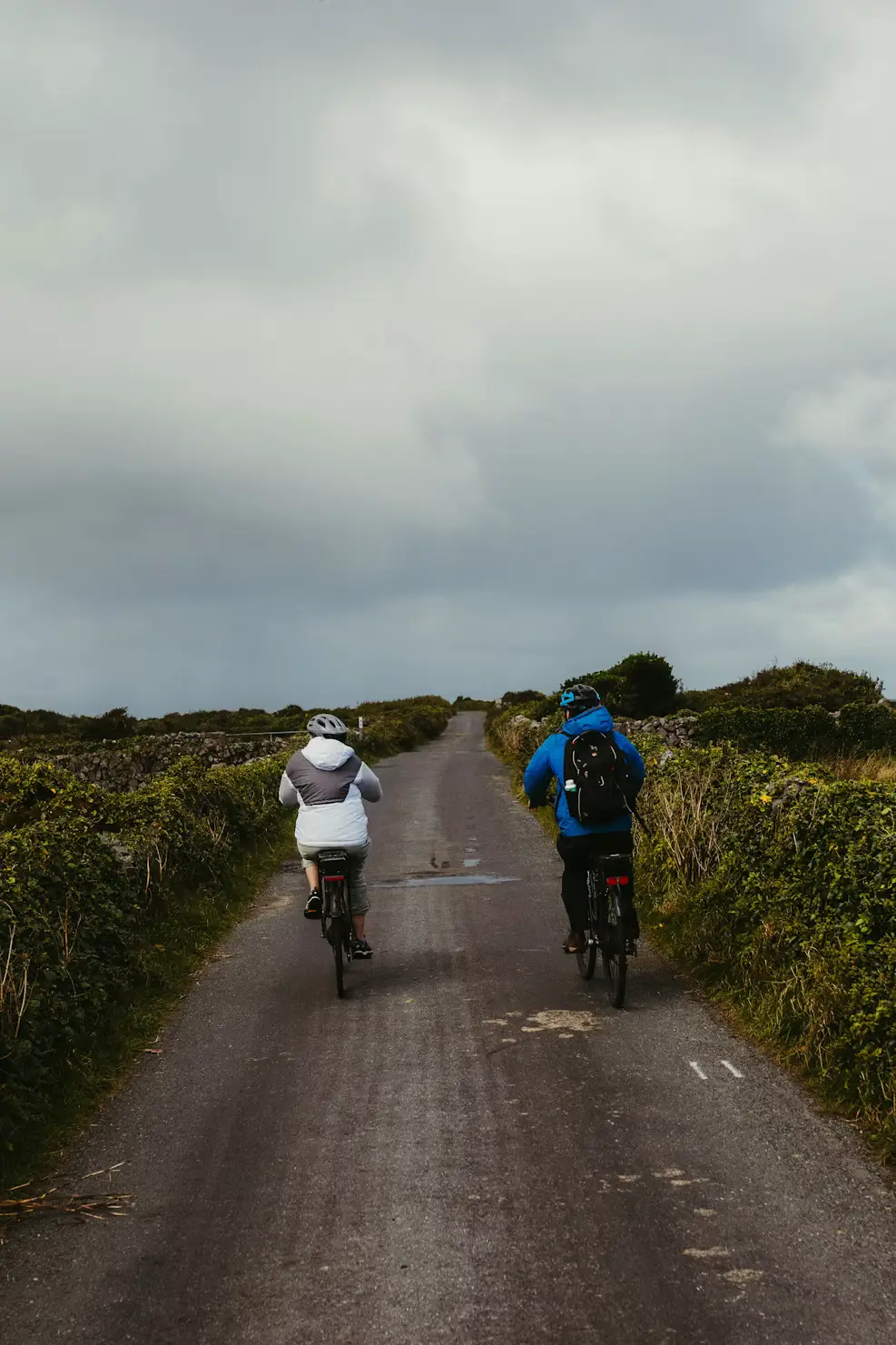 Two cyclists riding along a narrow country road bordered by hedges under a cloudy sky, Ireland.