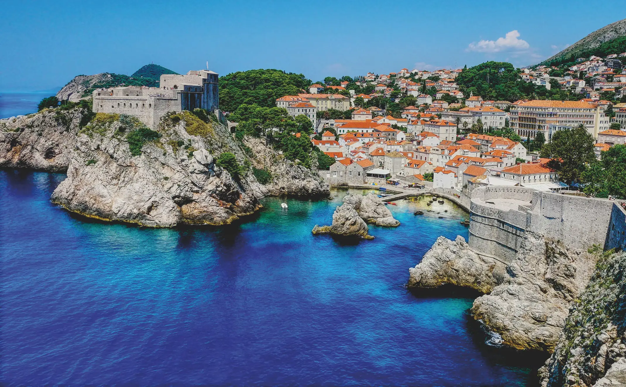 Rocky coastline near Dubrovnik with a stone fortress on a cliff overlooking deep blue water, red-roofed old town in the distance, Croatia.