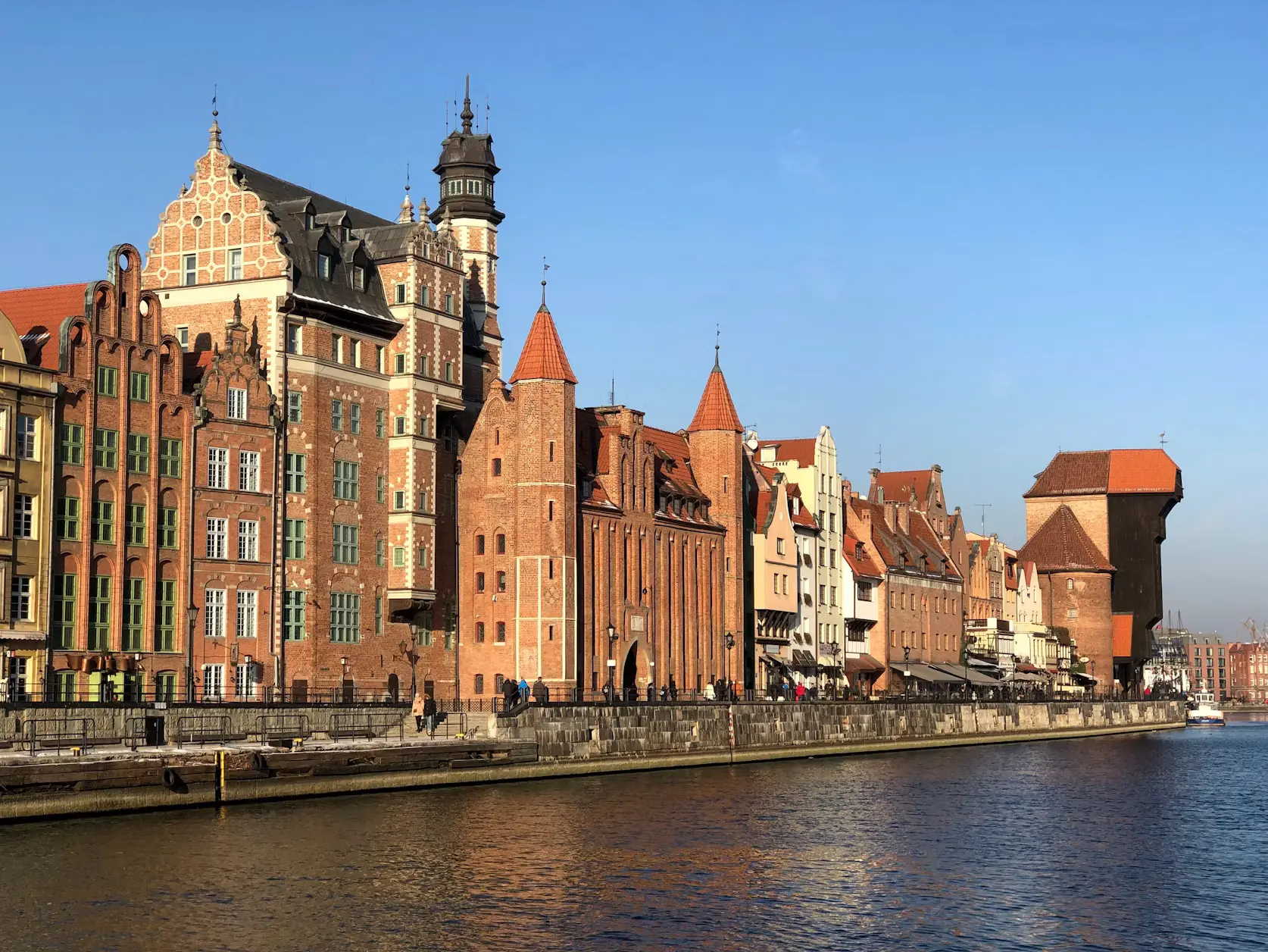Historic brick buildings and a wooden crane tower lining a riverside promenade under a clear blue sky, Gdańsk, Poland.