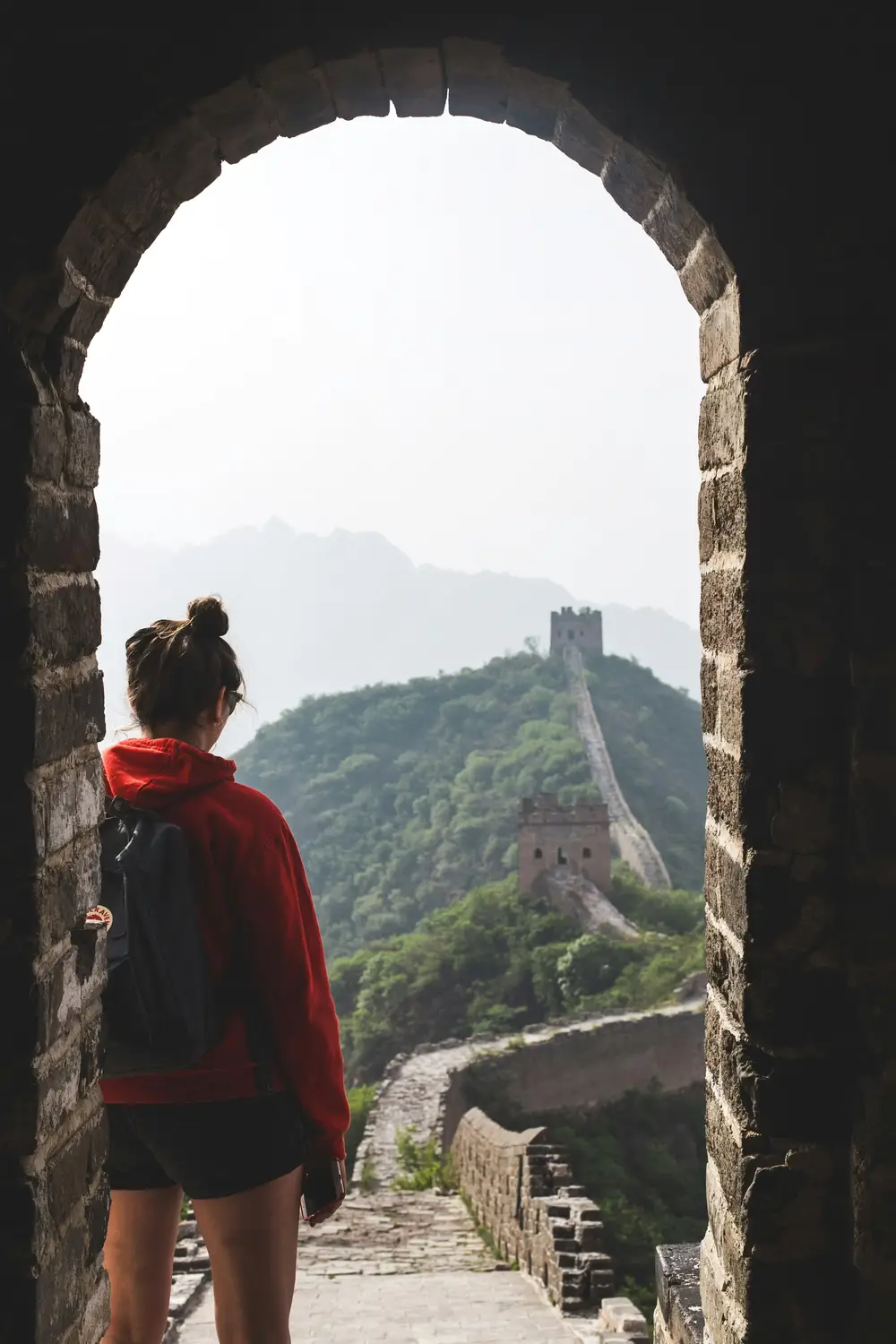 Woman in red hoodie looking through stone arch window of Great Wall of China watchtower toward misty mountains