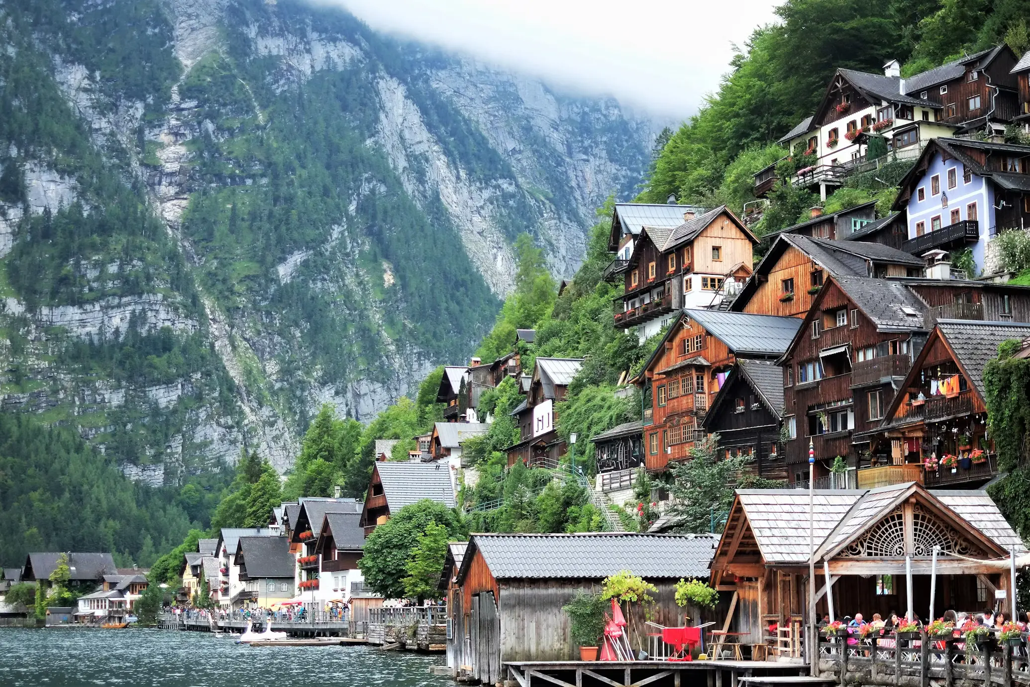 A lakeside alpine village with traditional wooden houses built into a steep, forested mountainside, with a boat dock and lakeside café in the foreground.