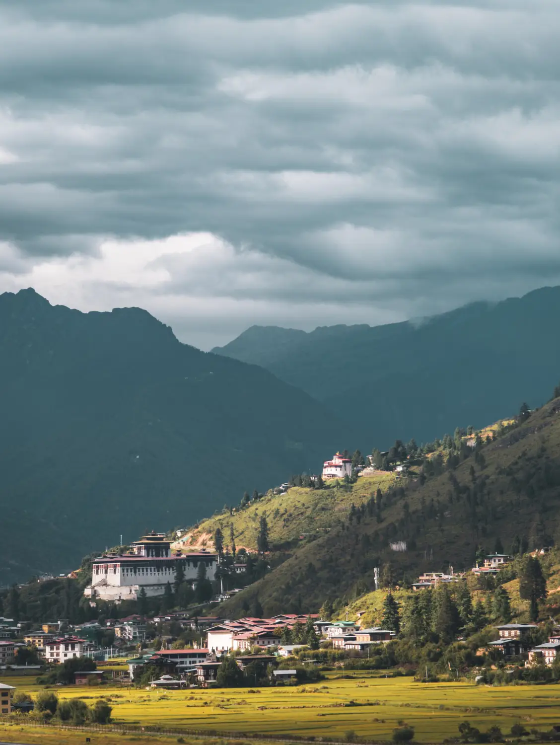 Aerial view of a valley town and dzong nestled among green fields beneath dramatic cloud-covered mountains in Bhutan.