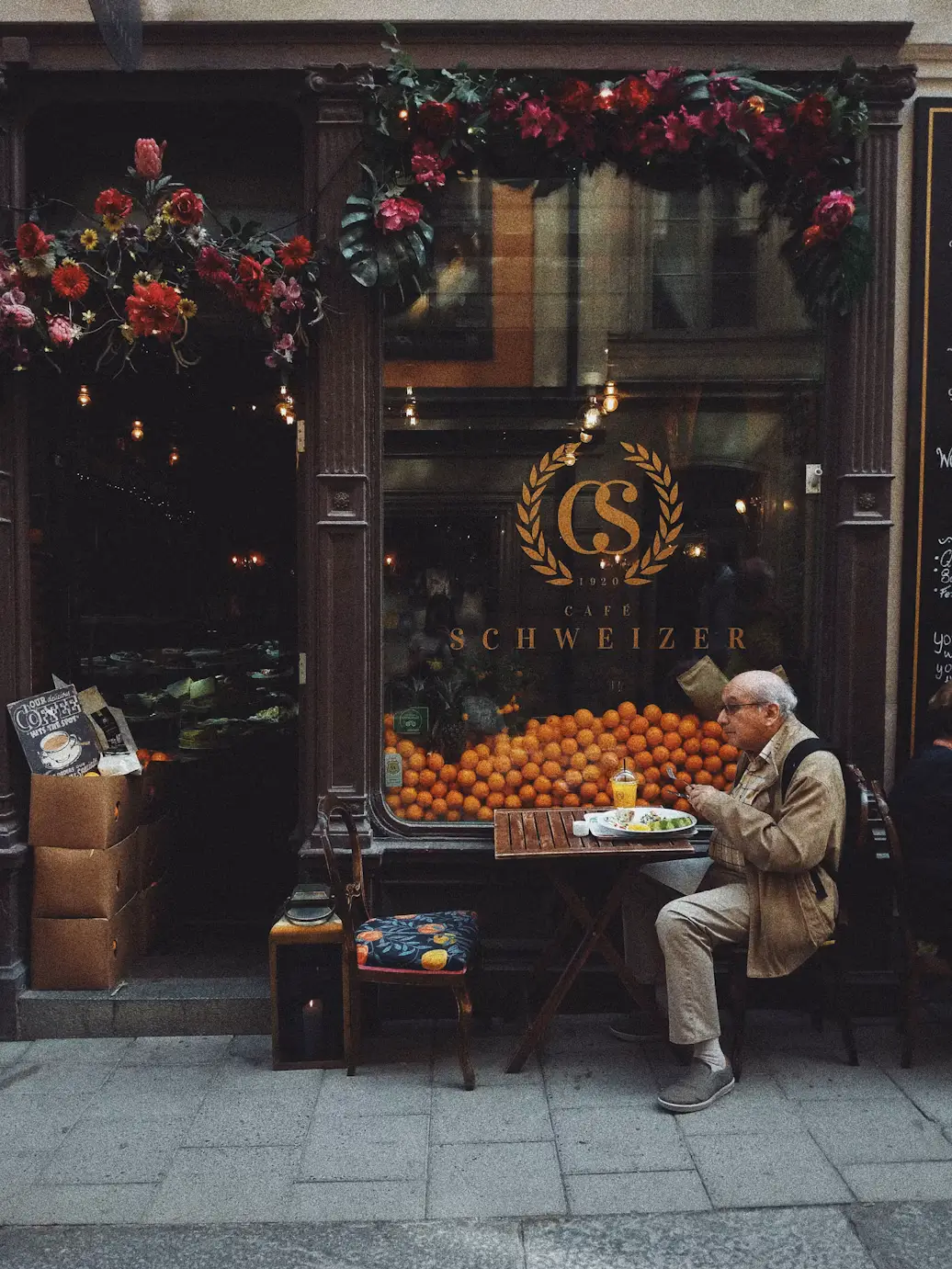 Elderly man seated outside a storefront with a fruit display, beneath a doorway decorated with hanging flowers.