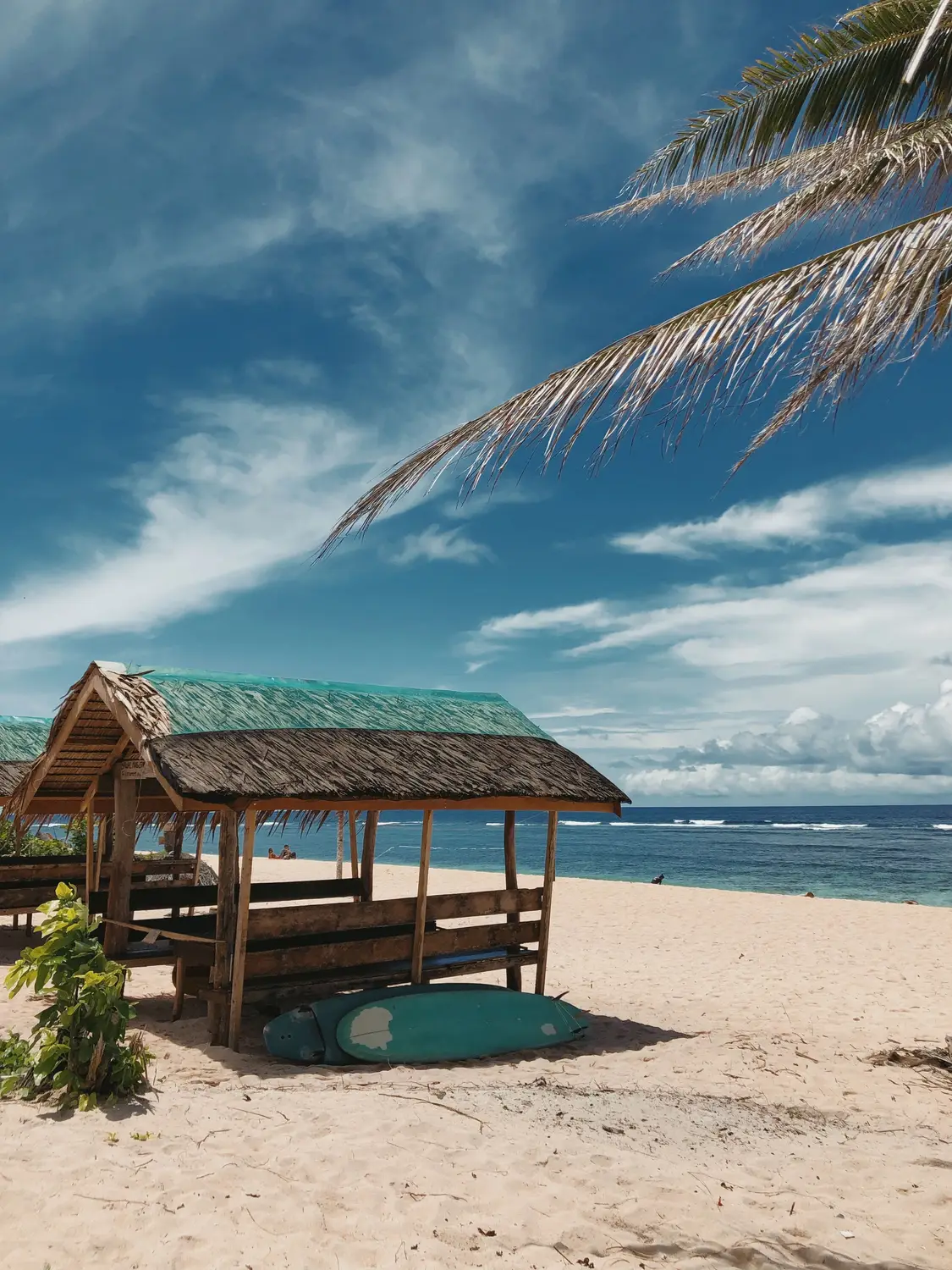 Wooden thatched beach hut with a surfboard on white sand beside turquoise sea and palm fronds in the Philippines.