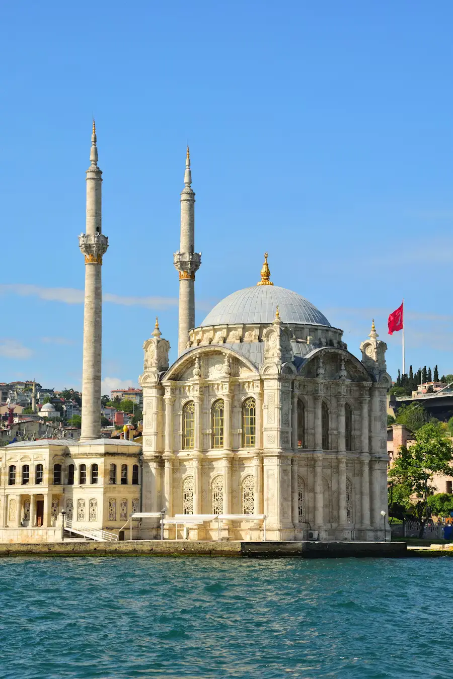 The white Ortaköy Mosque with twin minarets sitting on the shore of the Bosphorus in Istanbul, with a Turkish flag flying above.