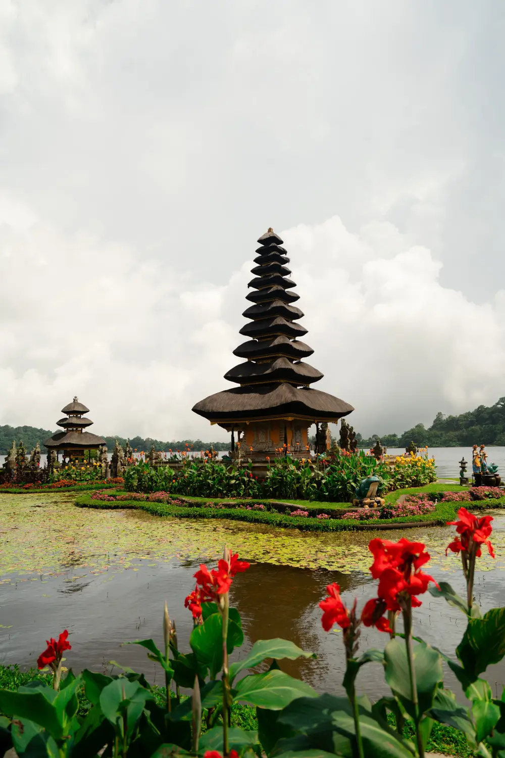 Pura Ulun Danu Beratan lake temple with tiered pagoda, framed by vibrant red canna flowers and manicured gardens in Bali, Indonesia.