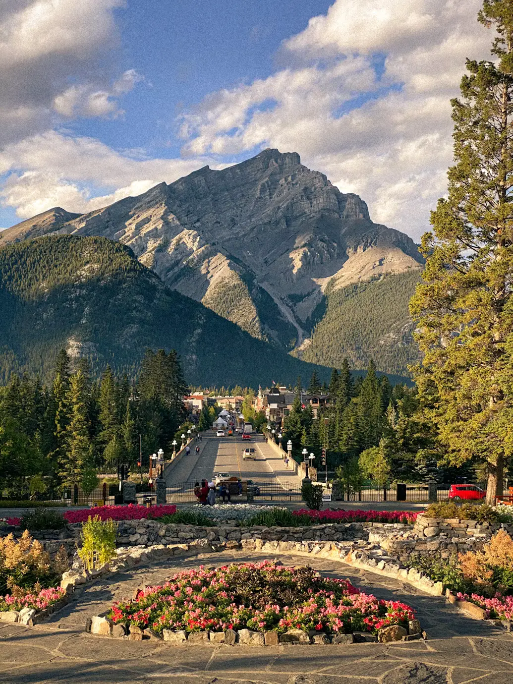 A flower-lined pathway leading toward a mountain peak, with pine trees and a partly cloudy sky in Banff.