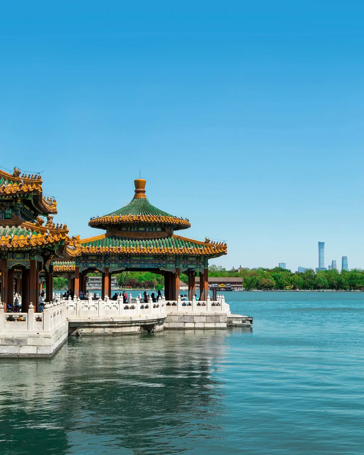 Traditional Chinese pavilion with green and gold glazed tile roof extending over Beihai Lake Beijing with modern skyline