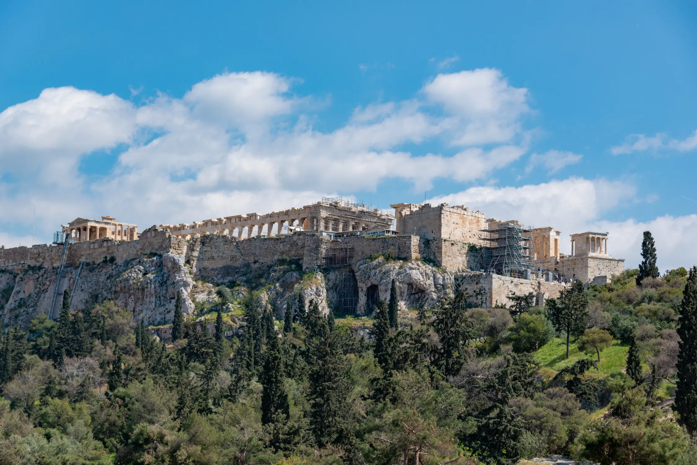 Ancient hilltop temple complex and stone ruins overlooking trees under a bright blue sky with scattered clouds.