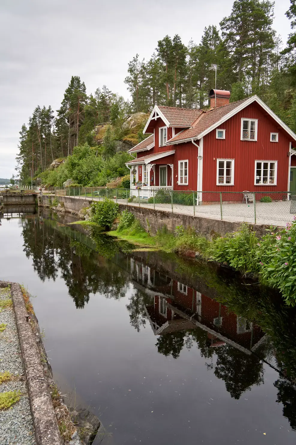 Red Swedish wooden houses on a wooded hillside, reflected in a calm canal below.
